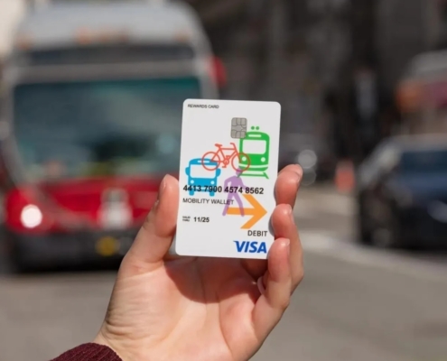 a hand holds a Mobility Wallet card as a bus approaches in the background
