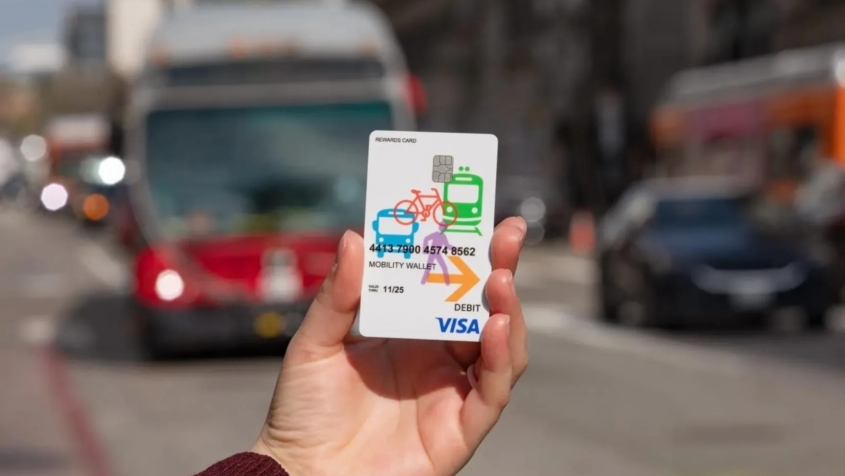 a hand holds a Mobility Wallet card as a bus approaches in the background