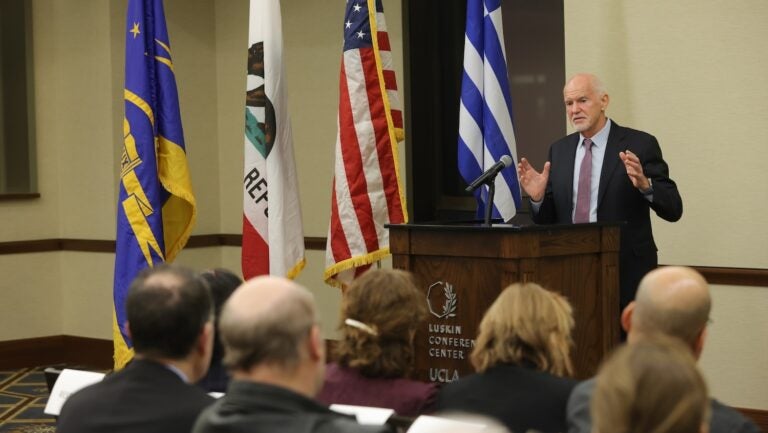 Former Greek prime minister George Papandreou speaking at UCLA. Greek flag and U.S. flag are behind him|woman with dark hair and glasses at podium