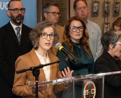 Woman in tan jacket at podium with others in background
