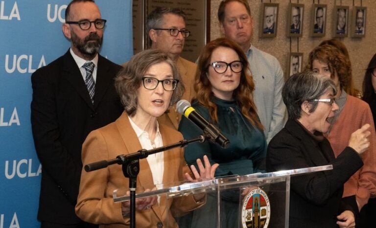 Woman in tan jacket at podium with others in background