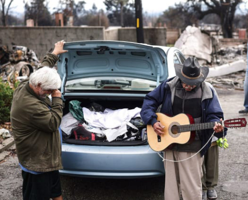 two people, one playing guitar, next to open car trunk amid fire zone