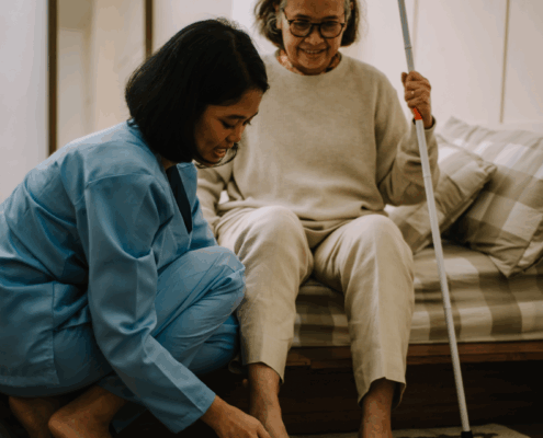 Female caretaker helps elderly woman put on her shoes beside the bed.