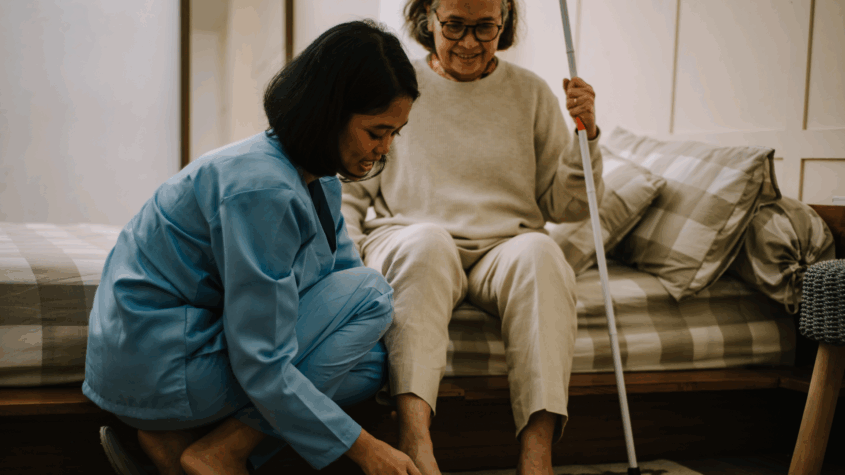 Female caretaker helps elderly woman put on her shoes beside the bed.