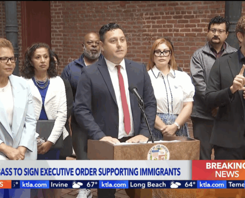 Mayor Karen Bass stands next to Edgar Garcia, MURP '06 who is at the pulpit introducing the mayor as she signs a new executive directive to protect immigrant communities amid ICE raids happening all across our city.