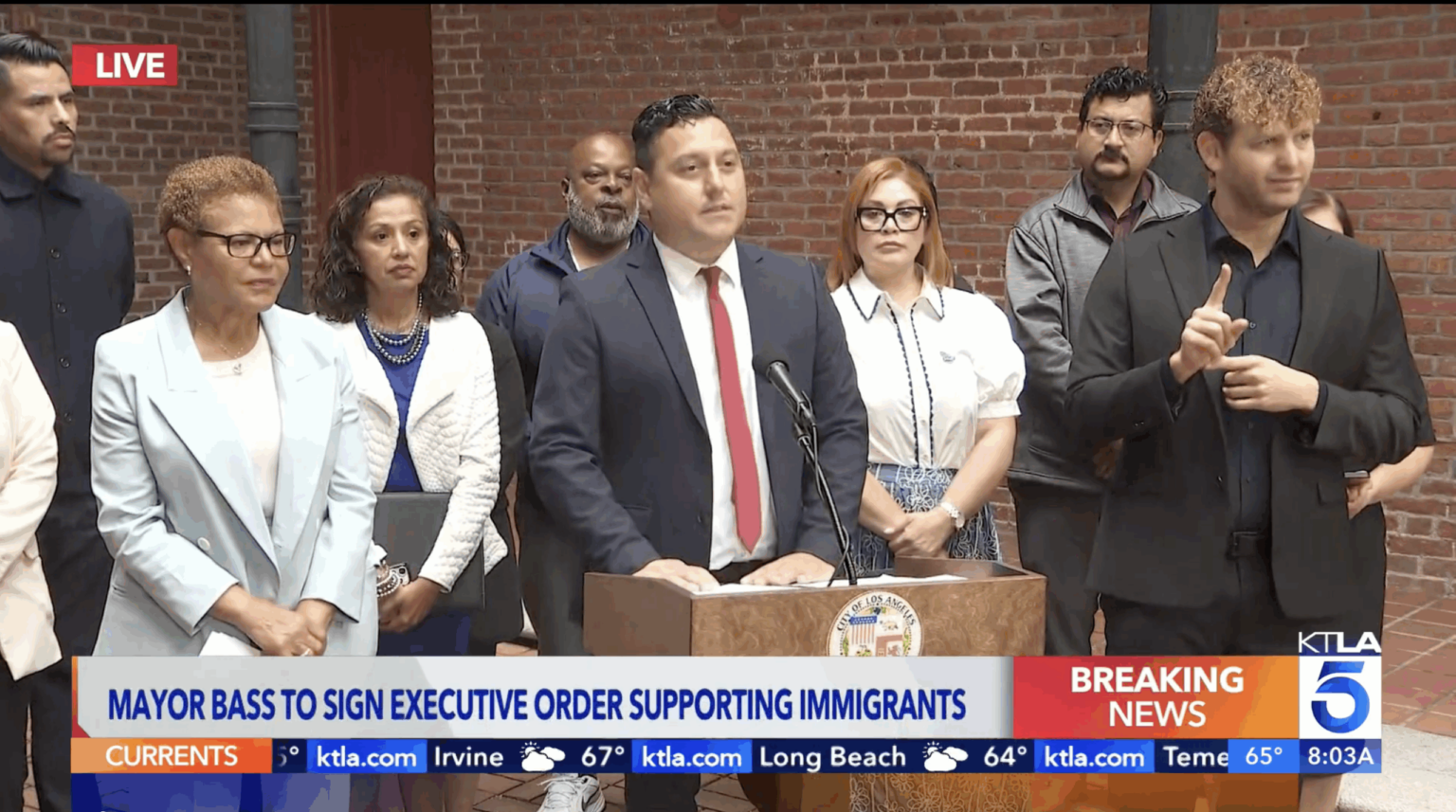 Mayor Karen Bass stands next to Edgar Garcia, MURP '06 who is at the pulpit introducing the mayor as she signs a new executive directive to protect immigrant communities amid ICE raids happening all across our city.||