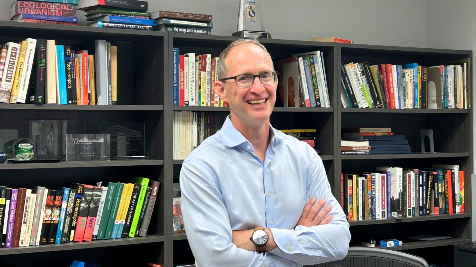 UCLA urban planning professor mike manville stands smiling in his office in front of a row of bookshelves.