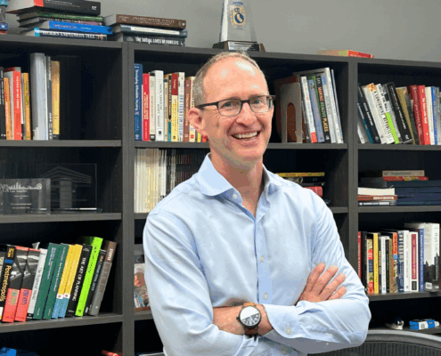 UCLA urban planning professor mike manville stands smiling in his office in front of a row of bookshelves.
