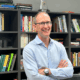 UCLA urban planning professor mike manville stands smiling in his office in front of a row of bookshelves.