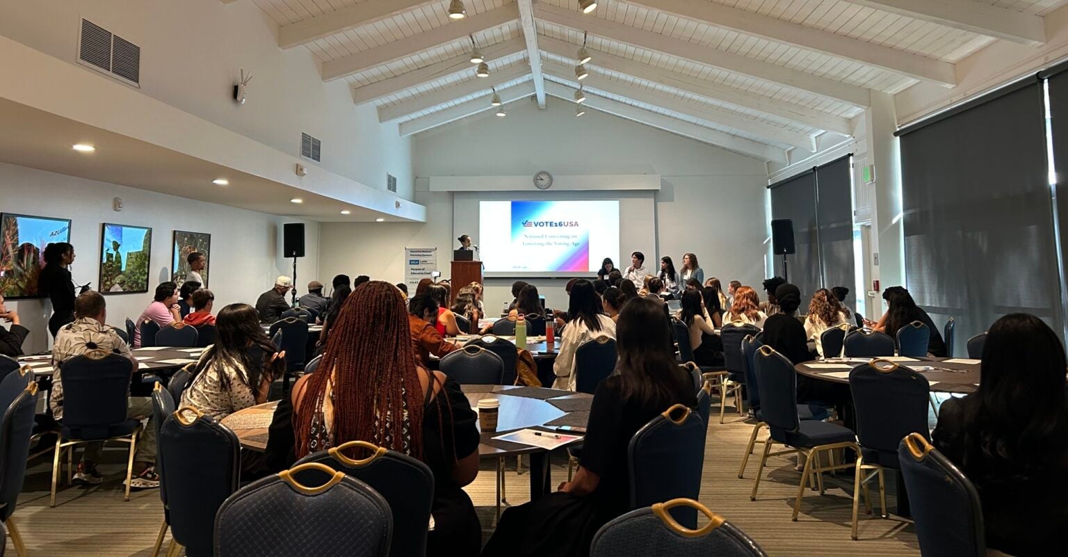 Large conference area with circle tables, dozens of youth seated while someone on stage does a presentation.