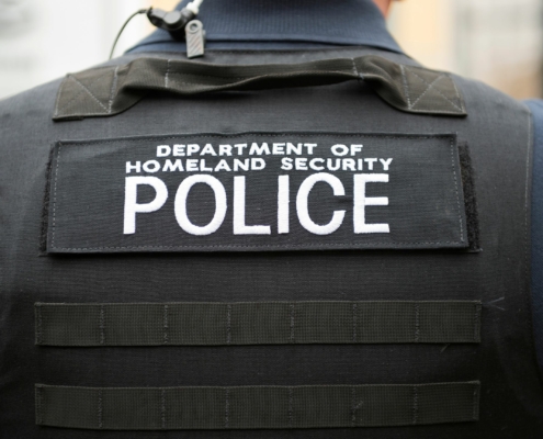Photo of a shoulder and back of a person wearing a bullet proof vest with the words Department of Homeland Security Police written across the back in white letters.