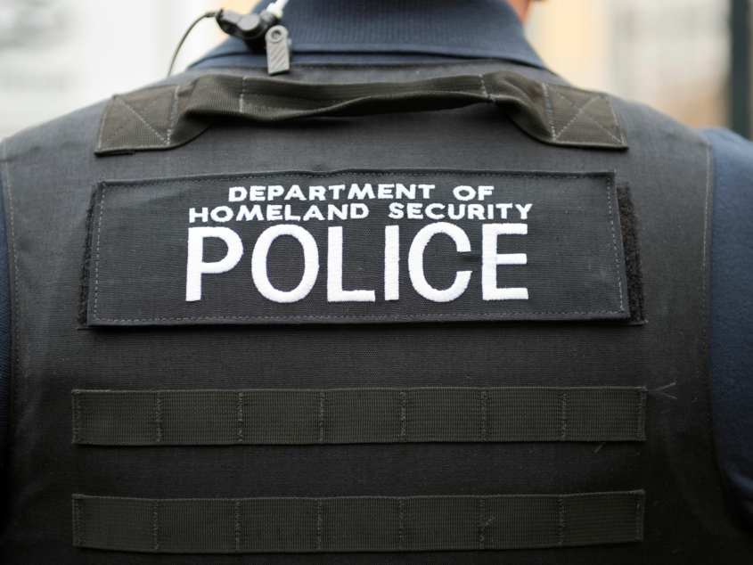 Photo of a shoulder and back of a person wearing a bullet proof vest with the words Department of Homeland Security Police written across the back in white letters.