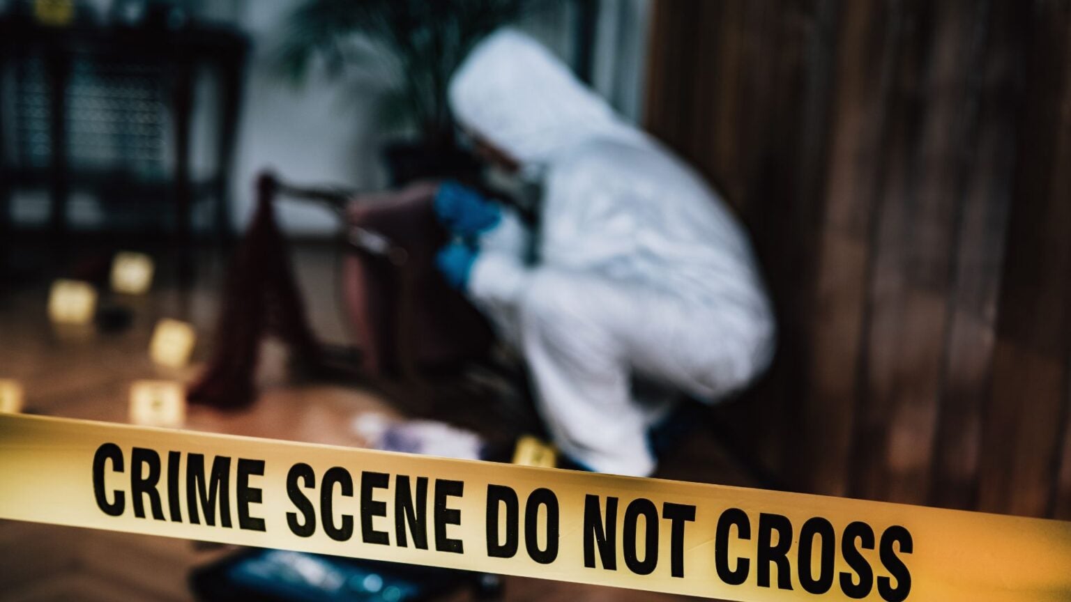 Crime scene yellow tape in the forefront with a detective in a white suit examining something on the ground in a forested area.