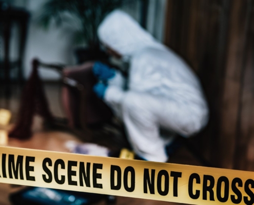 Crime scene yellow tape in the forefront with a detective in a white suit examining something on the ground in a forested area.