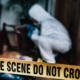 Crime scene yellow tape in the forefront with a detective in a white suit examining something on the ground in a forested area.