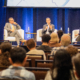 3 panelists speak in front of an audience on stage.