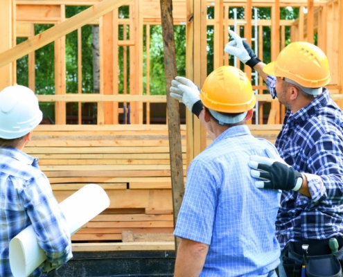 Three construction workers on a construction site with hard hats on pointing and talking together in front of a house with a wooden frame.