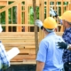 Three construction workers on a construction site with hard hats on pointing and talking together in front of a house with a wooden frame.