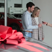 a new car at the dealership with a large red bow on the top, with three people in the background getting the keys to the brand new car.
