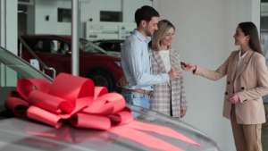 a new car at the dealership with a large red bow on the top, with three people in the background getting the keys to the brand new car.