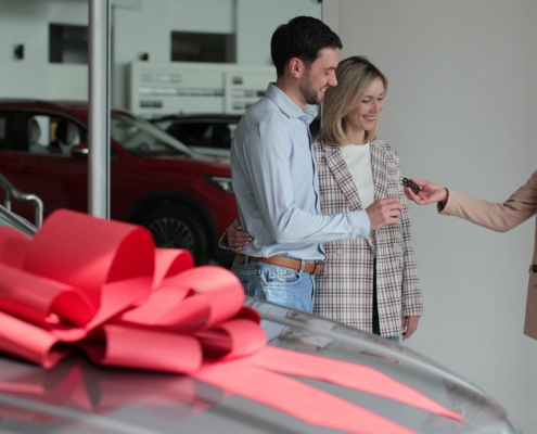 a new car at the dealership with a large red bow on the top, with three people in the background getting the keys to the brand new car.