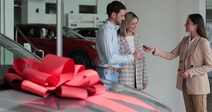 a new car at the dealership with a large red bow on the top, with three people in the background getting the keys to the brand new car.
