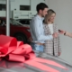 a new car at the dealership with a large red bow on the top, with three people in the background getting the keys to the brand new car.