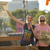 Sarah Jakle Speaking on a microphone at a political rally with a flyer that reads "politicians are no longer allowed to choose their voters"