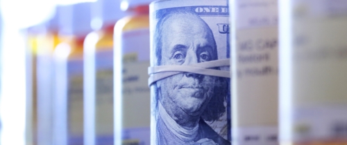 A roll of one hundred dollar bills sits among a row of prescription medication bottles.