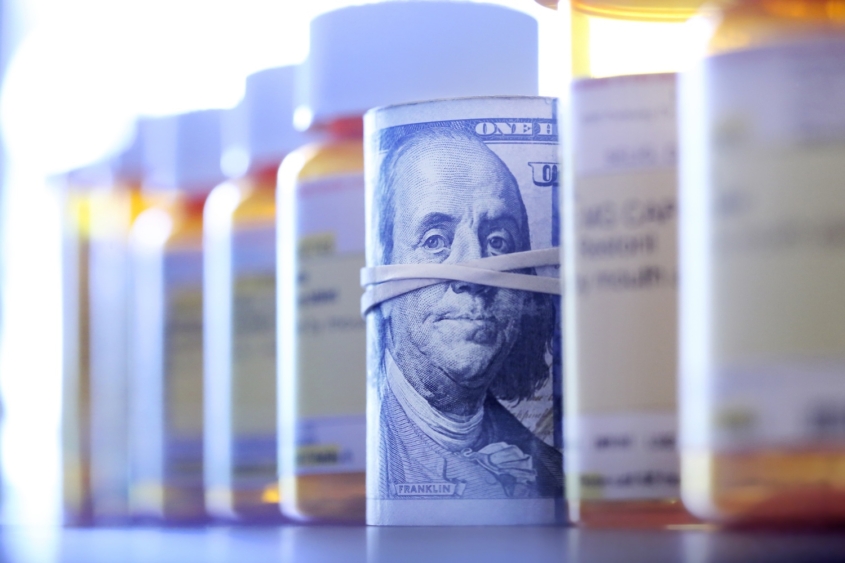 A roll of one hundred dollar bills sits among a row of prescription medication bottles.