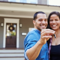 male and female holding up a key to their new home standing in front of the house they just purchased.