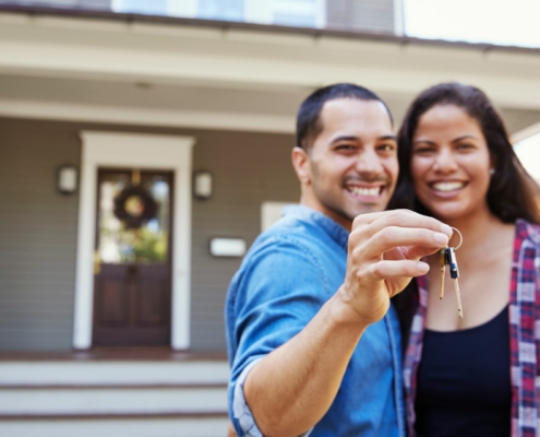 male and female holding up a key to their new home standing in front of the house they just purchased.