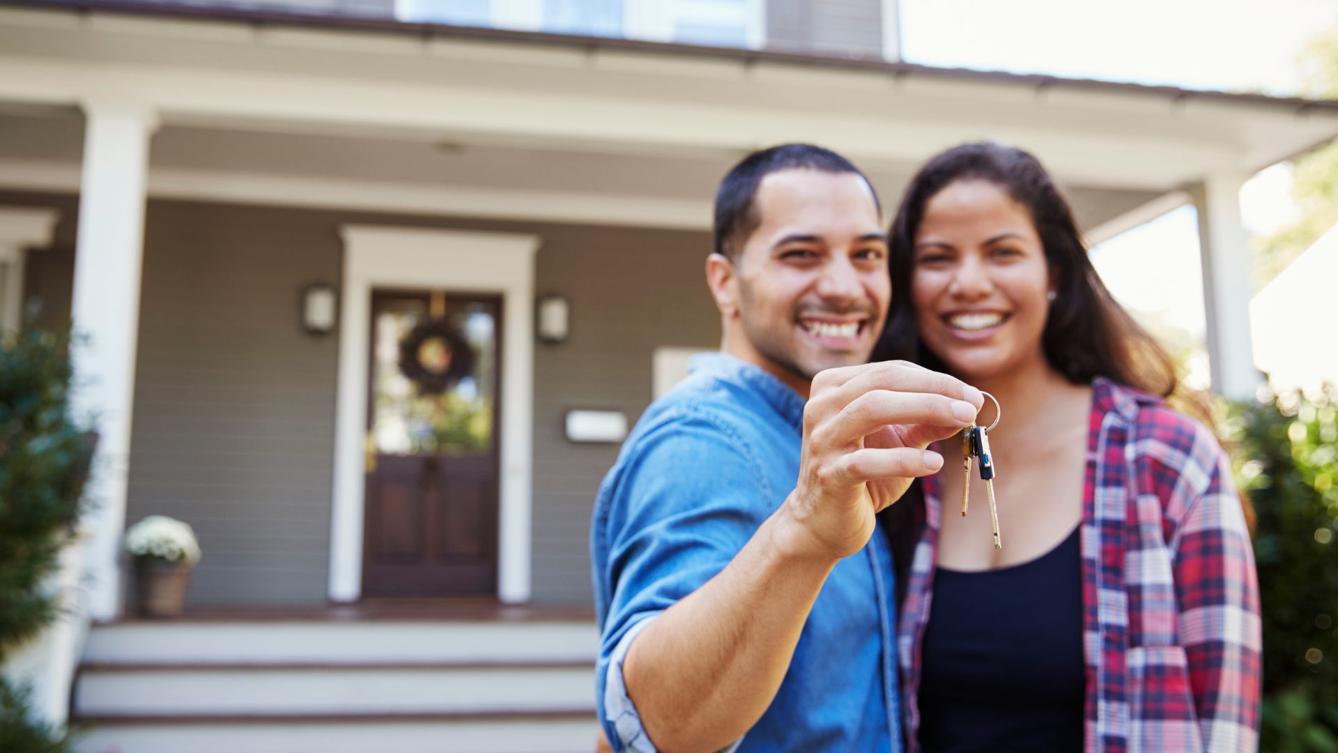 male and female holding up a key to their new home standing in front of the house they just purchased.
