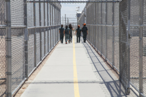 Group of people walking together in a fenced and gated ICE detention center.