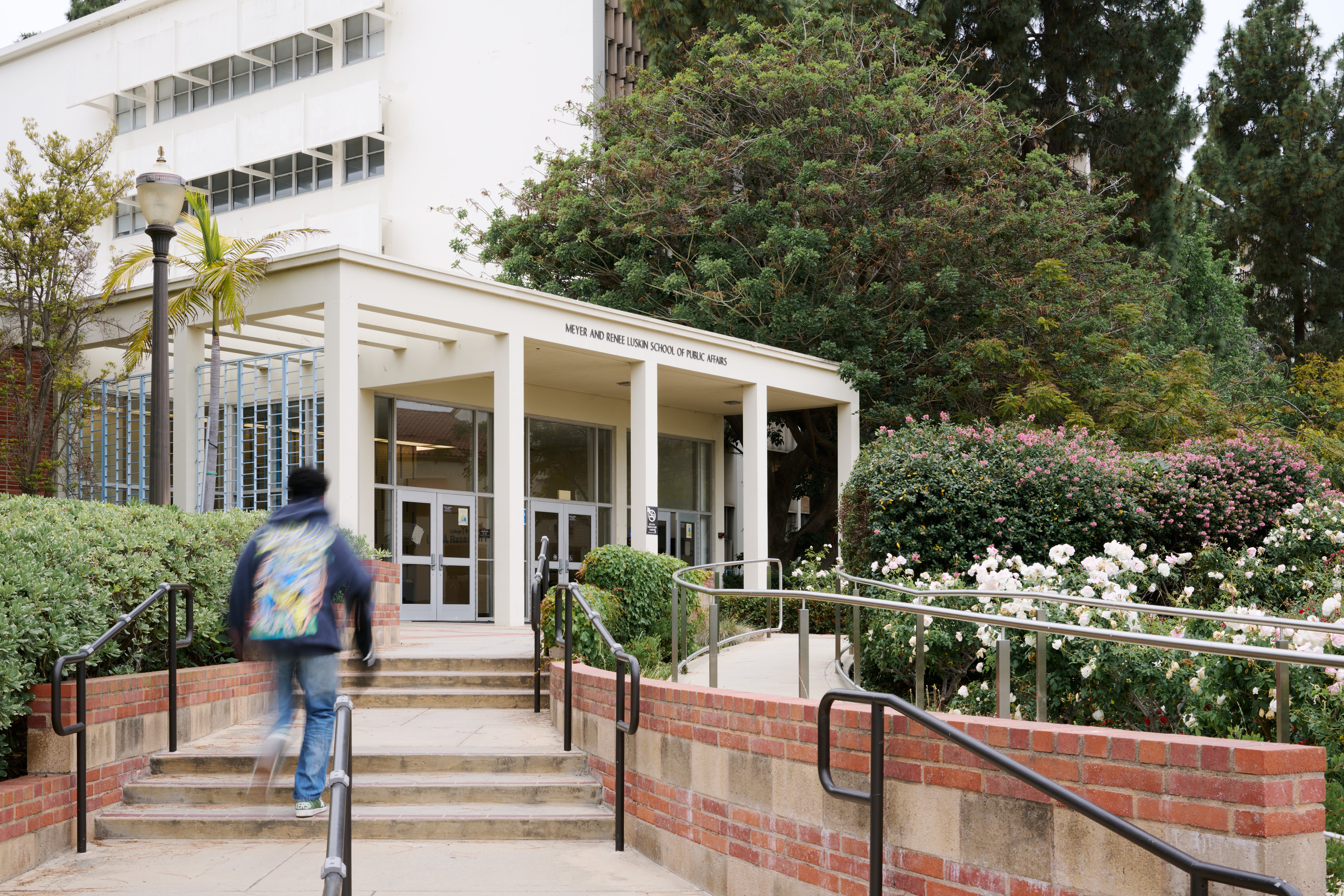 White building which is the east entrance of the UCLA Luskin school of public affairs building at UCLA with stairs leading up to the entrance, back of a student is walking up the stairs to the building entrance.