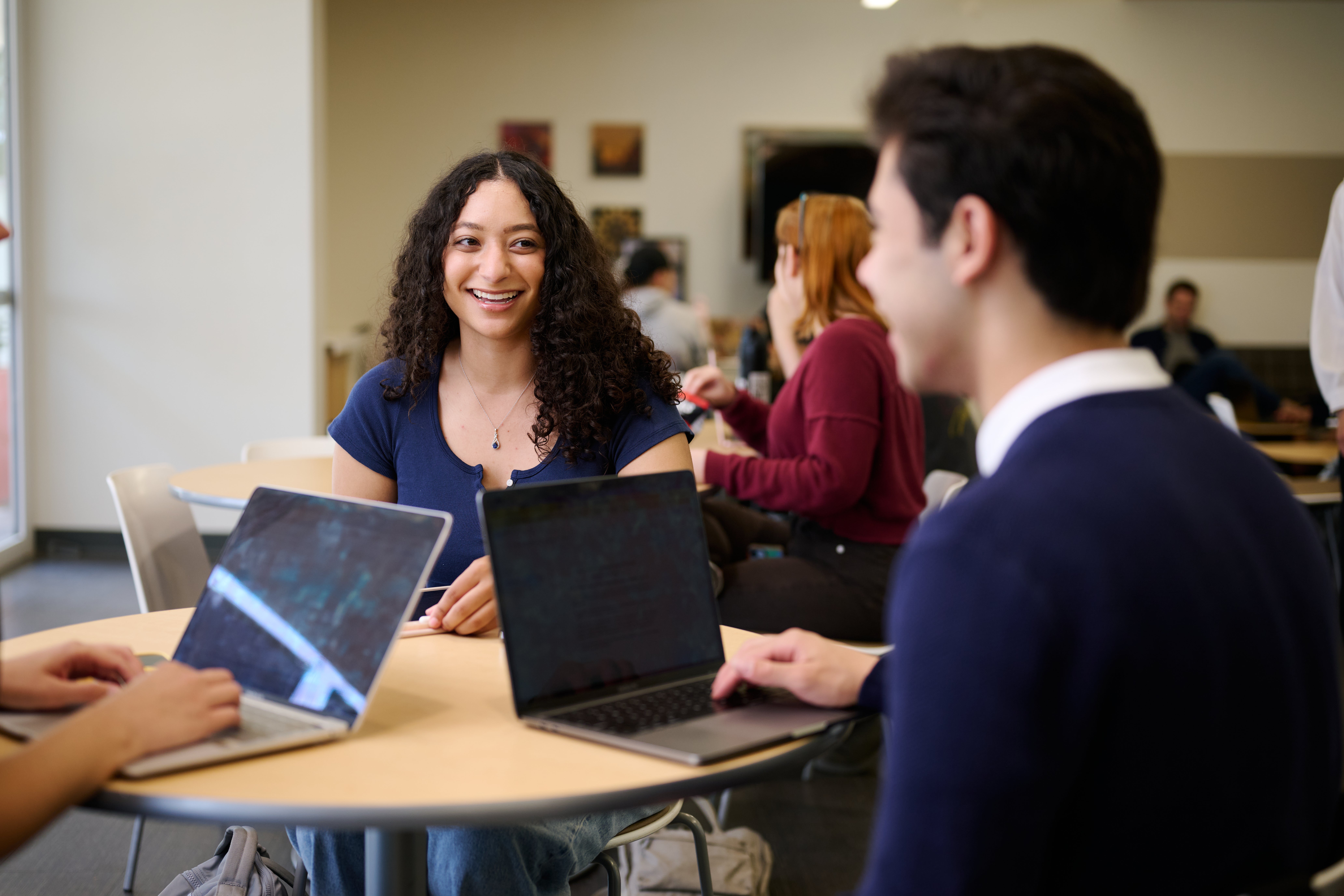 Luskin students sit around a table in the Luskin Commons smiling and talking with laptops on the table.