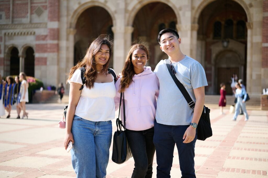 Three students stand shoulder to shoulder smiling at the camera in in front of Royce Hall on UCLA campus