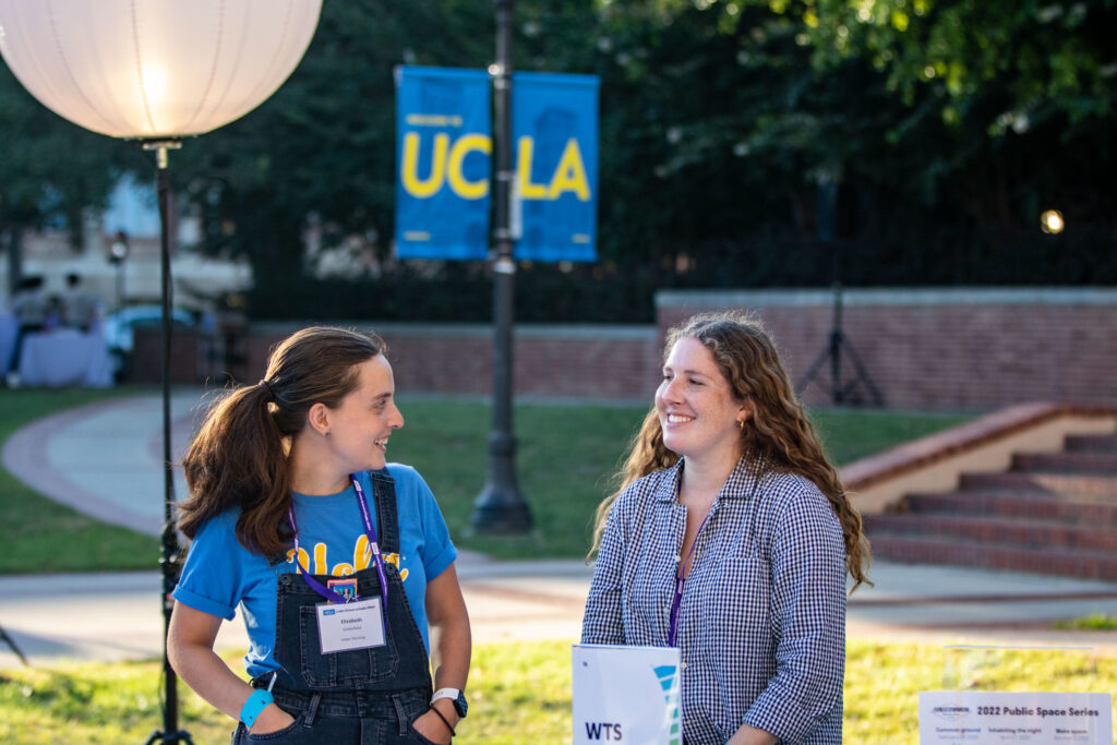 Two students at the annual Luskin Block Party standing in front of a UCLA sign, smiling smiling at each other on campus.