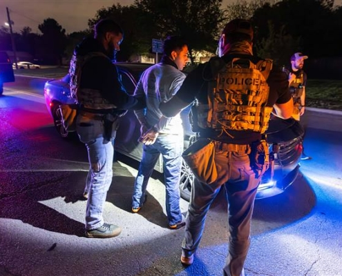 Immigration enforcement officers handcuff a man in the night, in front of a car in the middle of the road.