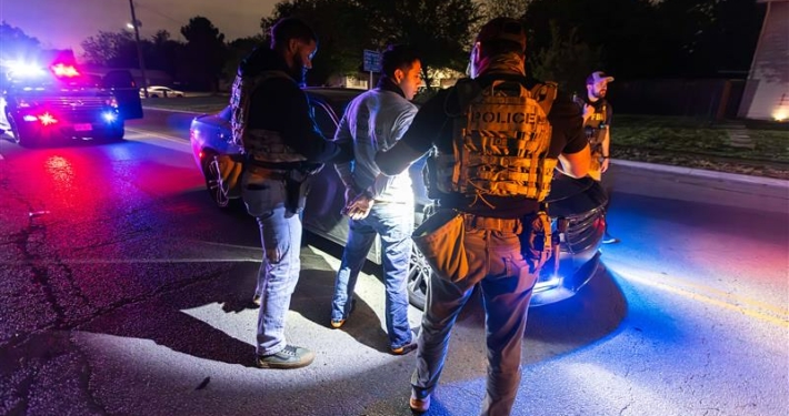Immigration enforcement officers handcuff a man in the night, in front of a car in the middle of the road.