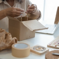 Rob Fairlie Entrepreneurship a woman who owns a jewelry small business is packaging a bracelet into a cardboard box on top of a table with lots of jewelry supplies and bags, she is fulfilling an online order.