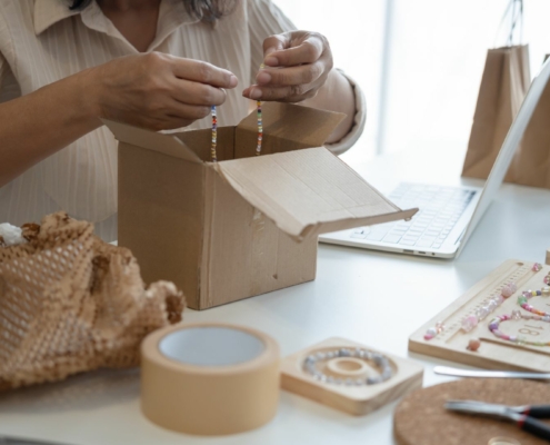a woman who owns a jewelry small business is packaging a bracelet into a cardboard box on top of a table with lots of jewelry supplies and bags, she is fulfilling an online order.