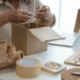 a woman who owns a jewelry small business is packaging a bracelet into a cardboard box on top of a table with lots of jewelry supplies and bags, she is fulfilling an online order.
