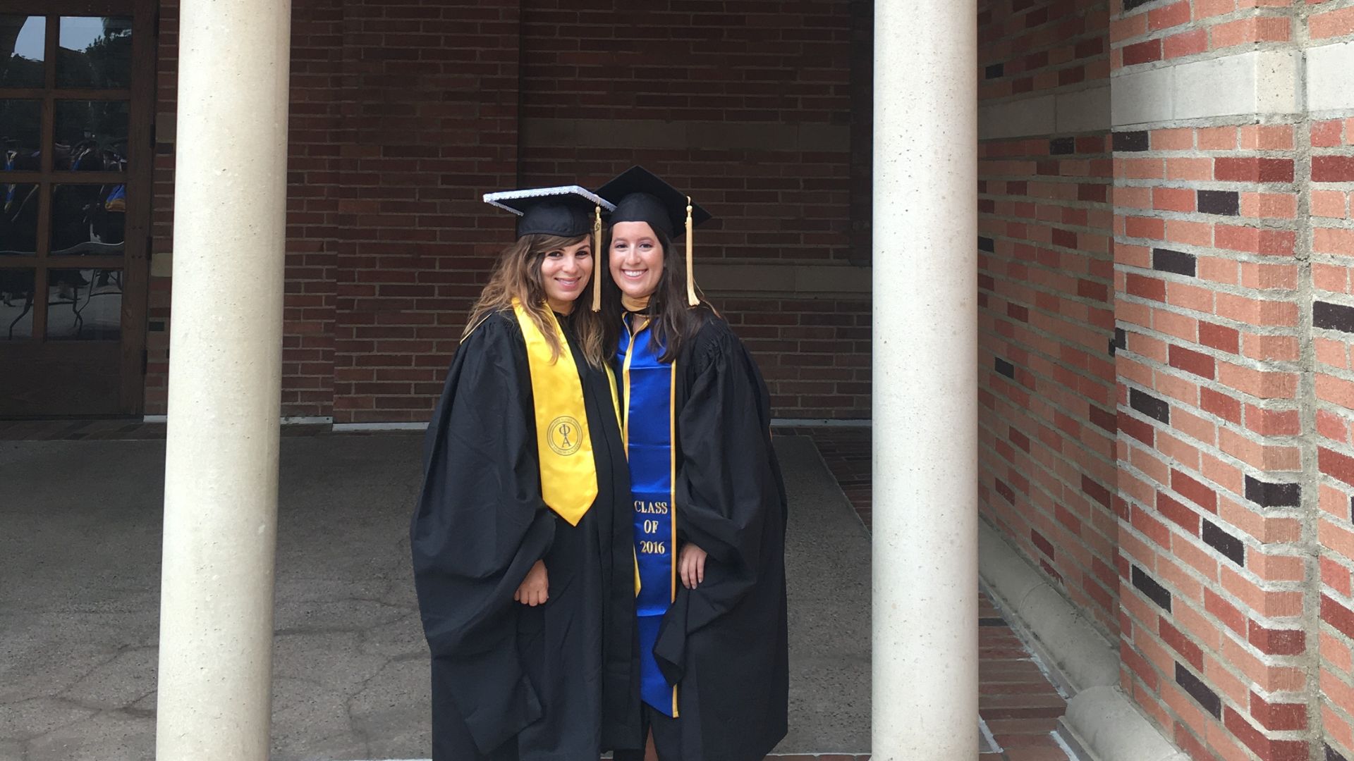 MSW Alumni Emily Dillon graduate of 2016 class, stands with her female friend, both are in cap and gown at UCLA graduation in front of a brick building.