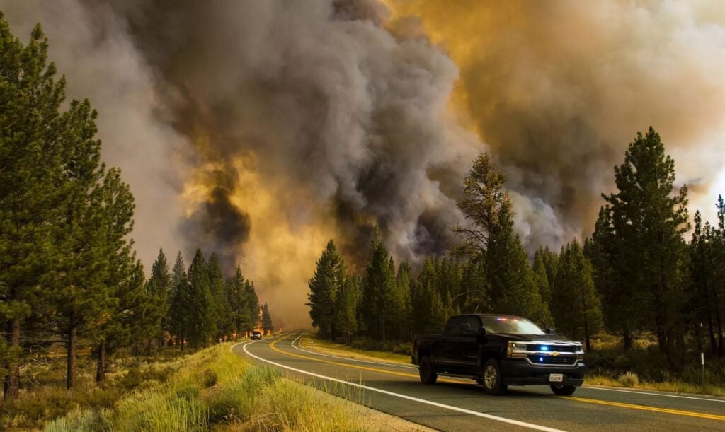 a car driving away from trees on fire and dark black smoke billowing in the sky