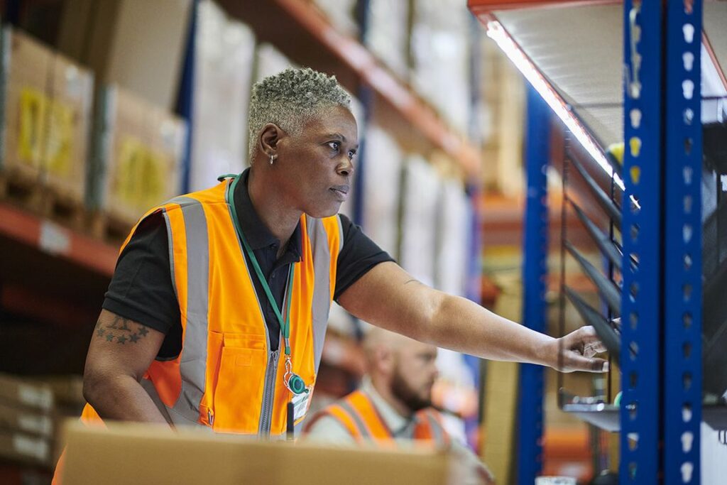 African American female warehouse worker wearing an orange vest doing her job.