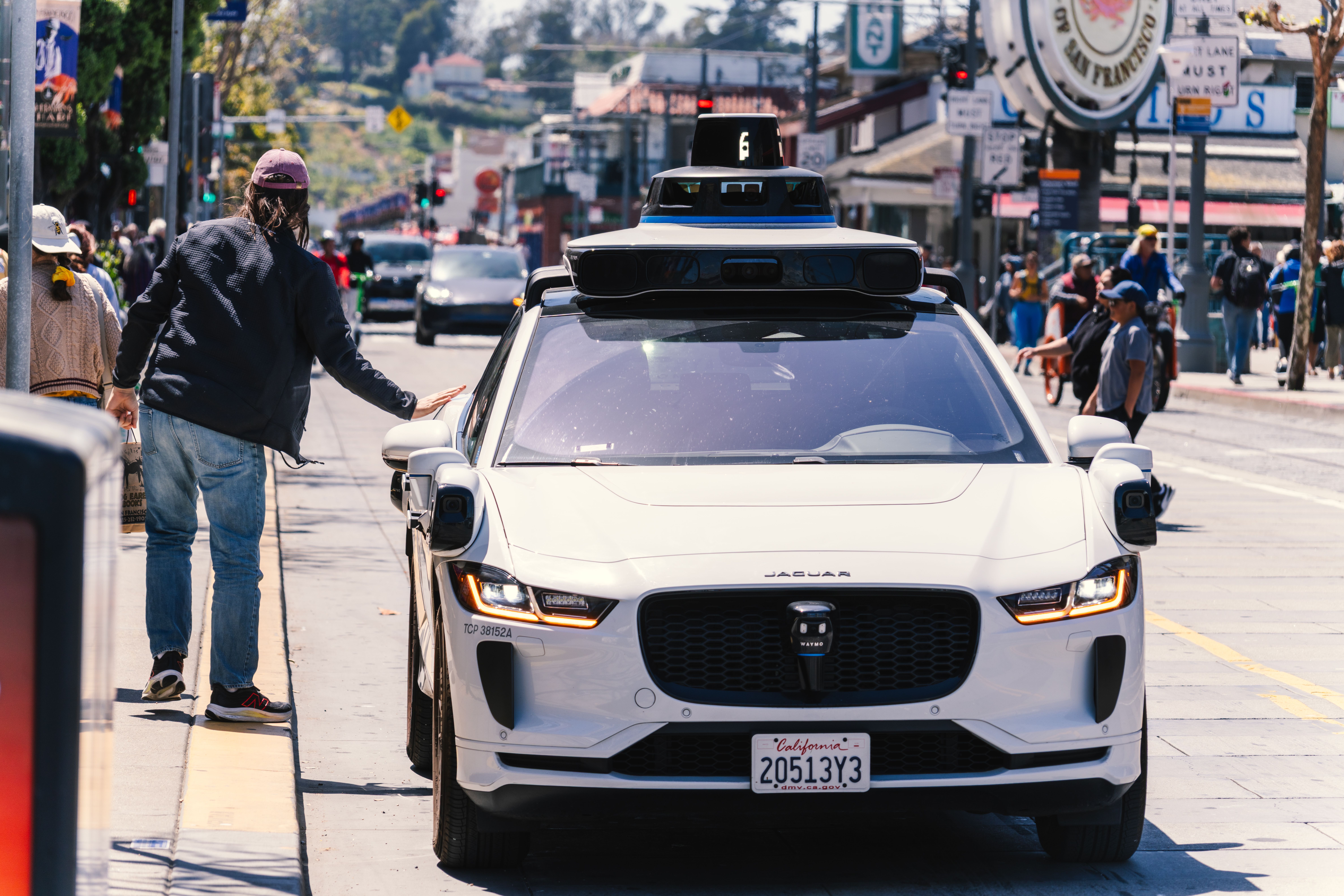 Waymo driverless taxi stops on the street while a passenger reaches out to open the door.