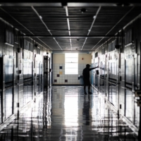 Dimly lit hallway of a high school, with lockers on either side, a man stands with his arms out touching a wall, conveying sadness.