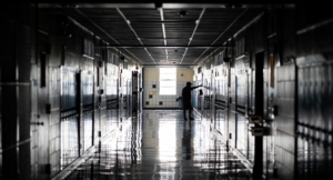 Dimly lit hallway of a high school, with lockers on either side, a man stands with his arms out touching a wall, conveying sadness.