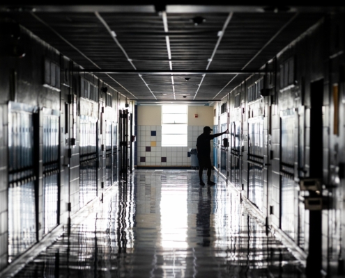 Dimly lit hallway of a high school, with lockers on either side, a man stands with his arms out touching a wall, conveying sadness.